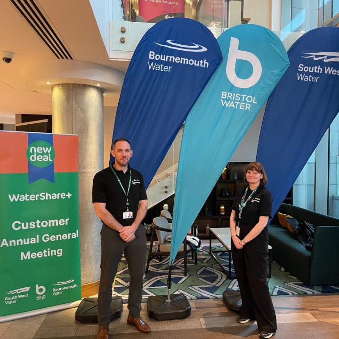Man and a woman stood next to branded flags and watershare roller banner sign